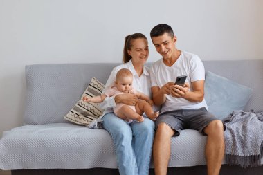 Olumlu genç bir aile, küçük kızlarıyla oturma odasında rahat bir öksürük içinde oturuyor. Babaları, karısına akıllı telefondan fotoğraflar gösteriyor. Olumlu duygular ifade ediyor..