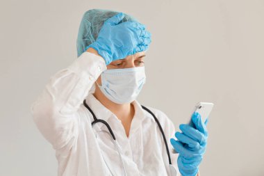 Depresyondaki genç kadın doktor tıbbi başlık, ameliyat maskesi, önlük ve lastik eldiven takıyor, hastanede çalışıyor, akıllı telefon kullanıyor, sorun yaşıyor ya da baş ağrısı çekiyor..