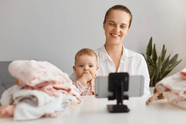穿着白衬衫 面带微笑的年轻女性和她可爱的小女儿一起在家里直播生活 为她的博客拍摄内容 看手机摄像头 — 图库照片