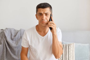 Üzgün, esmer, beyaz tişört giyen beyaz tenli yakışıklı bir adamın portresi öksürük üzerinde otururken kameraya bakıyor, dudak büküyor, kederini ifade ediyor..