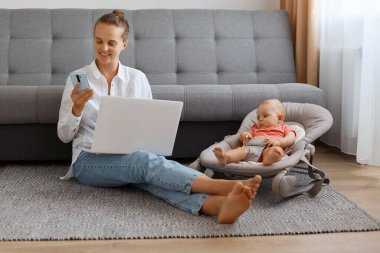 İyimser, beyaz tişörtlü, akıllı telefonu internette sörf yapmak için kullanan ve yeni doğmuş çocuğuyla fedai olarak vakit geçiren genç bir kadının yatay görüntüsü..