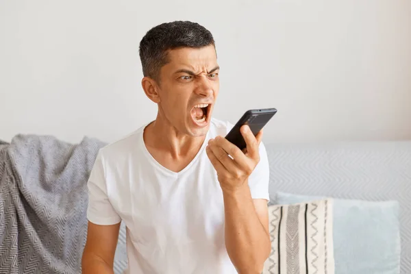 深褐色头发的年轻男性穿着白色休闲装坐在沙发上 手里拿着手机 怒气冲冲地尖叫着的横向照片 — 图库照片