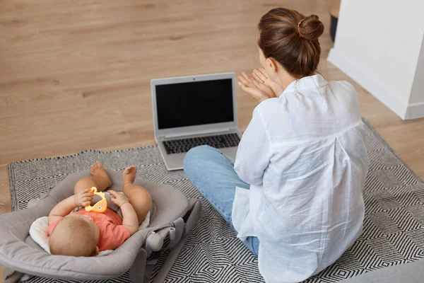バックビュー母の肖像画に座ってカーペットの近くのノートパソコンで彼女の幼児の娘でロッキングチェア 作業ノートブックで空のスクリーンのためのコピースペース — ストック写真