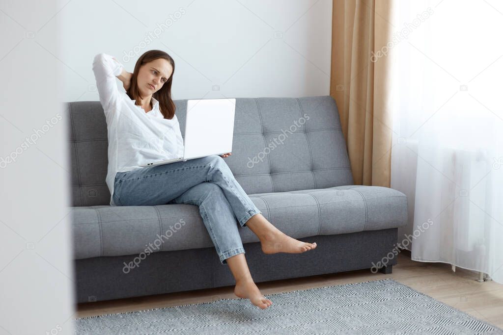 Tired young adult woman wearing white shirt and jeans sitting on sofa at home and working on laptop, looks exhausted, having pain in neck, long hours in front of computer.