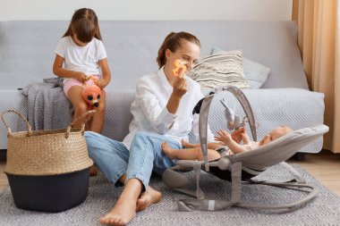 Bir kadın kızlarıyla evde vakit geçirir, anne bebeğiyle oynar, büyük kızı önemsemez, küçük kız başını eğip kanepede oturur ve kıskanır..