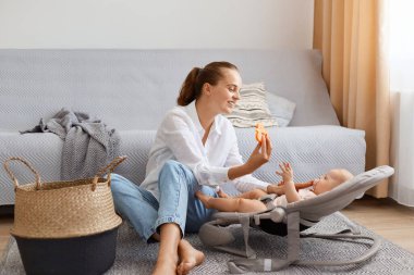 Beyaz gömlek ve kot pantolon giyen güzel bir kadının portresi öksürüğünün yanında oturuyor ve küçük bebeğiyle oynuyor, sallanan sandalyede kızına oyuncağını gösteriyor..