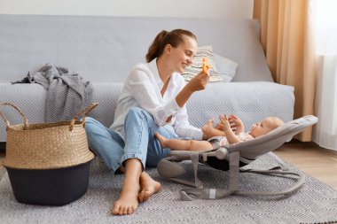 Evde genç bir anne ve şirin bir bebeğin evde yerde oynaması, sallanan sandalyede oturan kız ya da oğul, beyaz gömlek giyen anne ve çocuğa oyuncak gösteren kot pantolon giyen çocuk..
