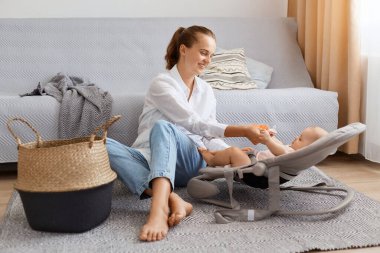 Tatmin olmuş yetişkin bir anne ve evde gri öksürüğün yanında yerde oynayan sevimli bir bebeğin uzun bir portresi. Anne, boş günlerinde kızıyla vakit geçiriyor..