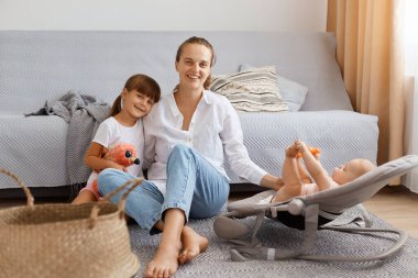 Anne ve küçük kızlar, bebek bakıcısı ve küçük çocuklar halının üzerinde oturan kız birlikte vakit geçirmenin tadını çıkarın, büyük çocuğa sarılan ve sallanan sandalyedeki küçük bebeğe dokunan kadın..