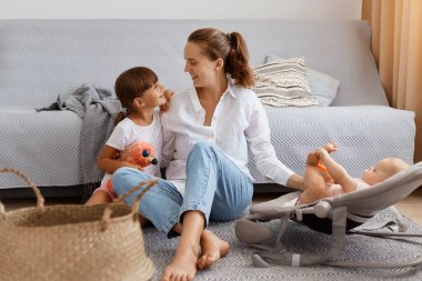 Beyaz gömlek ve kot pantolon giyen güzel bir kadın öksürüğünün yanında oturuyor, sevimli çocuklarıyla vakit geçiriyor, büyük kızına bakıyor ve sallanan sandalyede elini bebeğin bacağına koyuyor..