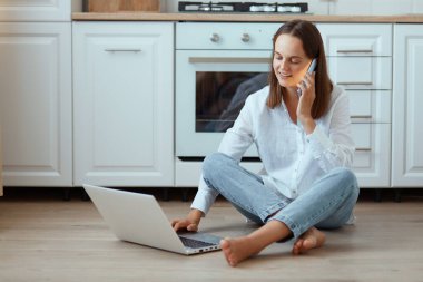 Yatay olarak gülümseyen, beyaz tişörtlü, kot pantolonlu bir kadının mutfakta oturmuş not defteri üzerinde çalışırken ve akıllı telefonla konuşurken pozitifliğini ifade ederken..