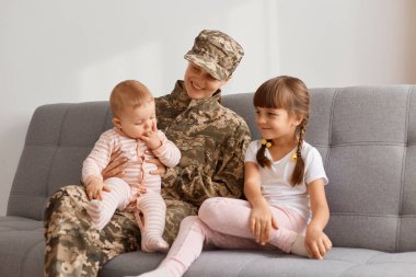 Kamuflaj üniforması giymiş, iki kızıyla birlikte kanepede oturan vatansever bir kadının gülümseyen bir fotoğrafı ordudan eve geldi, çocuklarını gördüğüne sevindi..