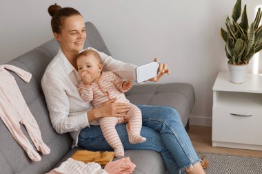 Beyaz gömlek ve kot giyen iyimser bir kadının profil portresi, yeni yürümeye başlayan kızıyla kanepede oturuyor ve aboneleriyle takılıyor, sosyal ağda canlı yayın yapıyor..
