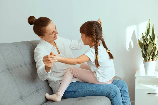 Indoor shot of happy smiling woman wearing white shirt and jeans, sitting on sofa with her charming daughter, family playing together, holding each others hands.