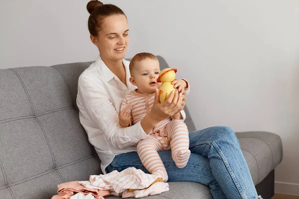 Beyaz gömlek ve kot pantolon giyen, küçük kızıyla kanepede oturan ve kız çocuğuna oyuncak gösteren, birlikte vakit geçiren beyaz tenli çekici bir kadın..