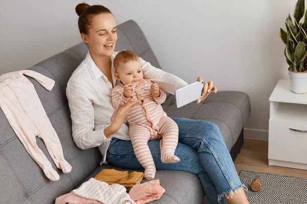 Beyaz gömlek ve kot pantolon giyen gülümseyen bir kadın, küçük kızıyla koltukta oturuyor, canlı yayın yapıyor, takipçileriyle konuşuyor, olumlu duygular ifade ediyor..