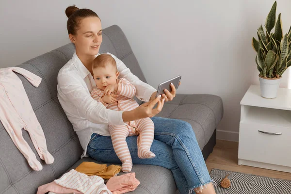 Beyaz kadın blogcu, koyu saçlı, koyu renk saçlı, günlük kıyafetler giyen, öksürük içinde bebeğiyle oturan ve annesi için video çeken ya da canlı yayın yapan biri..