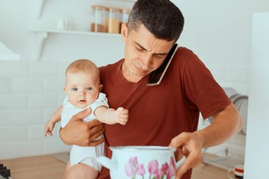 Koyu renk saçlı, beyaz, sıradan giyinen genç bir adamın yatay fotoğrafı. Bebek kızı tutan tişört, çorba ya da yulaf lapası taşıyan ot, telefonda konuşan, karısına kızını nasıl besleyeceğini soran..