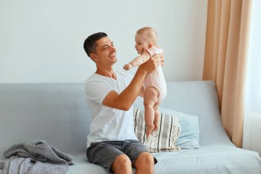 Beyaz t-shirt ve kot pantolon giyen yakışıklı bir adamın küçük kızıyla pencerenin yanındaki koltukta oturup mutlu bir şekilde poz verişini..