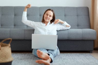 Beyaz gömlek ve kot pantolon giyen keyifli bir kadın dizüstü bilgisayarda çalışıyor, işini bitiriyor, ellerini uzatıyor, uzun saatler çalıştıktan sonra rahatlamaya çalışıyor..