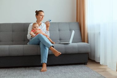 Beyaz tişörtlü, kot pantolonlu, üzgün bir kadının bebek kızıyla koltukta oturduğu, doğum izni sırasında internetten telefon açtığı bir fotoğraf..