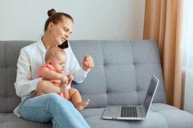 Beyaz gömlek ve kot pantolon giyen, küçük kızıyla koltukta oturan, arkadaşıyla veya ortağıyla cep telefonuyla konuşan, bilgisayar ekranına bakan çekici, koyu renk saçlı bir kadının kapalı çekimleri..