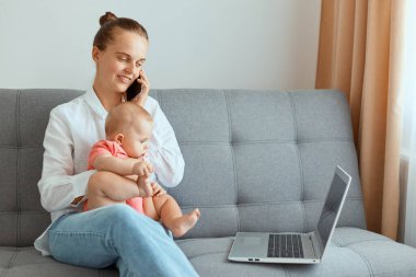 Beyaz gömlek ve kot pantolon giyen, bebek kızıyla koltukta oturan, cep telefonuyla konuşan ve dizüstü bilgisayarla çalışan tatmin olmuş bir kadının yatay görüntüsü..