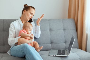 Kafası karışmış, beyaz tişörtlü, kot pantolonlu, doğum izni sırasında internetten çalışan, partneriyle telefonda konuşan, bebek kızıyla kanepede oturan kafası karışık kadının kapalı bir fotoğrafı..