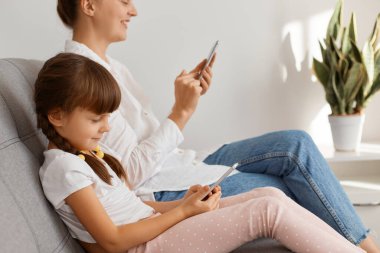 Beyaz gömlek ve kot pantolon giyip arka planda oturan annesiyle akıllı telefondan oyun oynayan sevimli küçük kız. İnsanlar olumlu duygular ifade ediyor. İnternette ailece vakit geçiriyorlar..