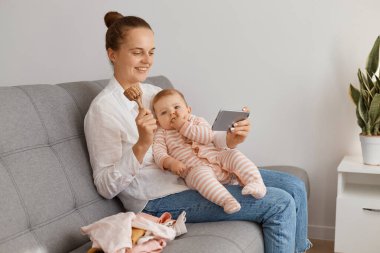 Tişört ve kot pantolon giyen, küçük kızıyla koltukta oturan ve canlı yayın yapan, takipçilere ahşap oyuncakları gösteren, cep telefonuyla video görüşmesi yapan pozitif bir kadın..