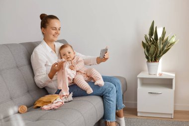 Bun saç stili olan beyaz gömlekli ve kotlu bir kadın blogcunun profil portresi. Yeni yürümeye başlayan çocuğuyla öksürük üzerinde oturuyor. Annesinin vlog 'u için yeni video kaydediyor..