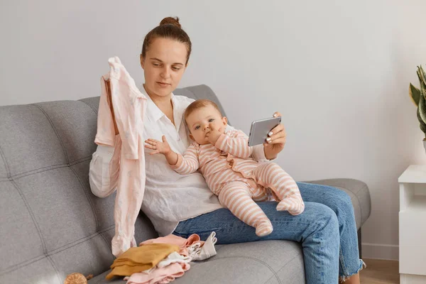 Beyaz gömlek ve kot pantolon giyen, öksürük içinde oturan, akıllı telefon ve kızının yeni kıyafetlerini elinde tutan kadın blogcu..
