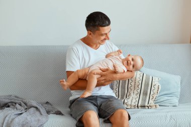 Beyaz t-shirt ve kot pantolon giyen yakışıklı esmer adamın elinde bebekle kanepeye oturup tatlı kızına baktığı ve çocuğuna tek başına baktığı bir fotoğraf..