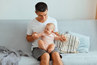 Beyaz tişört ve kot pantolon giyen yakışıklı, siyah saçlı bir adamın öksürük içinde kucağında bebek, mutlu bir baba ve çocukluk fotoğrafı..