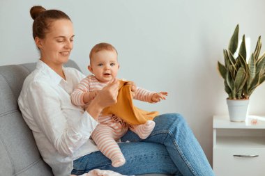 Küçük kızıyla birlikte kanepede oturan ve elinde yeni giysiler olan güzel, genç bir annenin mutlu ve güzel çocuğuna yeni giysiler aldığı yatay bir fotoğraf..