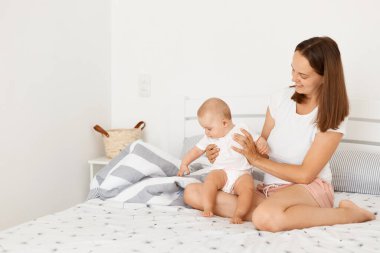 Beyaz tişörtlü çekici bir kadının bebek bebekle yatak odasında poz verdiği, annesinin kızını kucağında tuttuğu, olumlu duygular ifade ettiği kapalı çekimler..