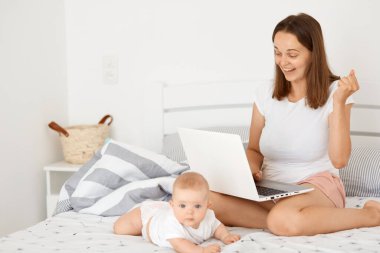 Son derece mutlu, koyu renk saçlı, beyaz tişörtlü bir kadın, bebeğiyle yatakta oturuyor ve internette çalışıyor, taşınabilir defterini tutuyor, yumruğunu sıkıyor, online bahis oynuyor ve kazanıyor..