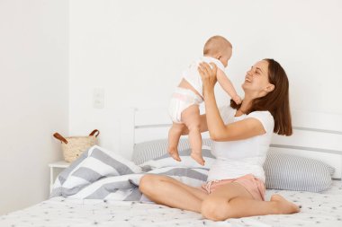 Çekici, mutlu, beyaz tişörtlü bir anne. Bebek bebekle yatak odasında oturuyor. Bir kadın çocuğunu kucağında tutuyor ve gülümsüyor..