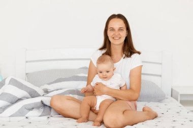 Olumlu iyimser anne beyaz bir tişört giyer, bebek bebekle yatak odasında oturur, dişlek gülümsemeyle kameraya bakar, pozitif duygularını ifade eder..