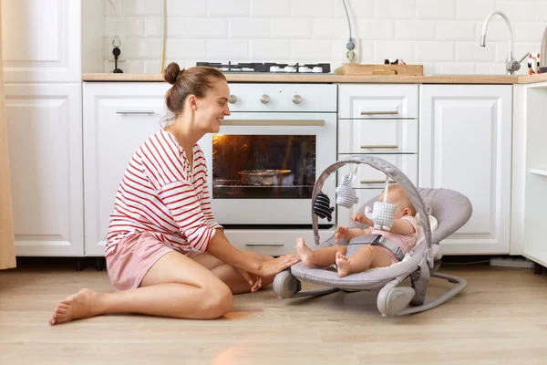Annenin yeni doğmuş kızıyla sallanan sandalyede turta pişirirken, günlük kıyafet giyen genç bir kadın ve beyaz bir mutfakta yemek pişirirken çekilmiş bir fotoğraf..