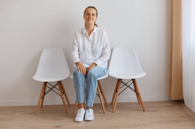 Gülümseyen, tatmin olmuş, beyaz duvara yaslanmış, gömlek ve kot giyen, sakin ve iyimser bir yüz ifadesiyle kameraya bakan genç ve çekici bir kadın..