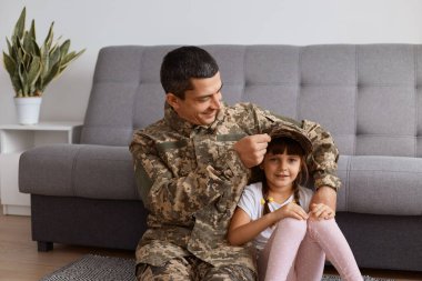 Kapalı alanda, asker üniforması giyen genç bir adam ordudan eve dönerken, kızıyla vakit geçirirken, şapkasını çocuğun kafasına takarken, mutlu bir şekilde gülümserken..