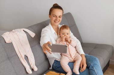 Kapalı alanda, beyaz tişörtlü ve kotlu mutlu bir kadın bebeğiyle öksürüyor, video görüşmesi yapıyor, cep telefonunu tutuyor, büyüleyici bir gülümsemeyle cihaza bakıyor..