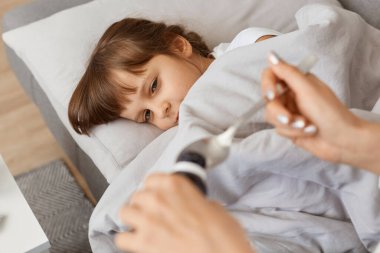 Küçük hasta kızın kanepede yattığı, annesinin kaşığını şurupla verdiği, baş ağrısı ve yüksek ateş çeken ve grip belirtileri gösteren bir fotoğraf..