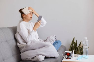 Bun saçlı, beyaz kazak giyen, battaniyeye sarılı, elinde telefonla oturan ve grip belirtileri gösteren hasta kadın portresi..