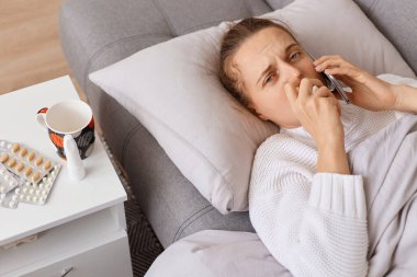 Hasta kadının kanepede yatıp telefonla konuşurken, kameraya bakarken, yüksek ateşi, burnu akarken ve baş ağrısı çekerken, tedaviyi sormak için doktoru ararken..