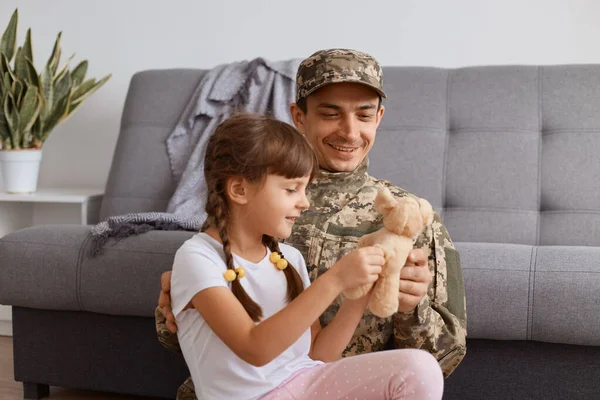Horizontal shot of delighted father with his kid, Caucasian soldier man wearing camouflage uniform returning home after army, enjoying time with his little daughter, child showing her toy to dad.
