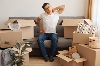 Beyaz tişört ve kot pantolon giyen yorgun hasta bir adamın portresi. Etrafı paket yığınlarıyla çevrili, boynuna masaj yapan, yeni bir yere taşınmaktan bıkmış..