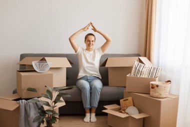 Pozitif iyimser kadın beyaz t-shirt ve kot pantolon giyerek yeni bir evde ya da dairede oturuyor, ellerini başının üstünde tutuyor, kollarıyla çatı yapıyor, evinde güvende..