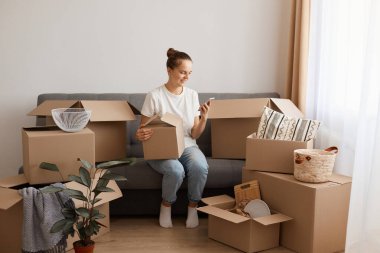 Beyaz tişört ve kot pantolon giyen beyaz bir kadının taşındıktan sonra yeni bir evde ya da dairede oturduğu, eşyalarla dolu bir kutuyu tuttuğu ve akıllı telefon kullandığı, mesaj yazdığı kapalı mekan fotoğrafı..